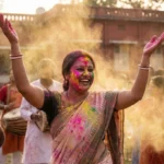 A joyous woman, covered in colorful powder, celebrates with raised arms amidst a cloud of yellow dust during a vibrant festival, with others playing musical instruments in the background.