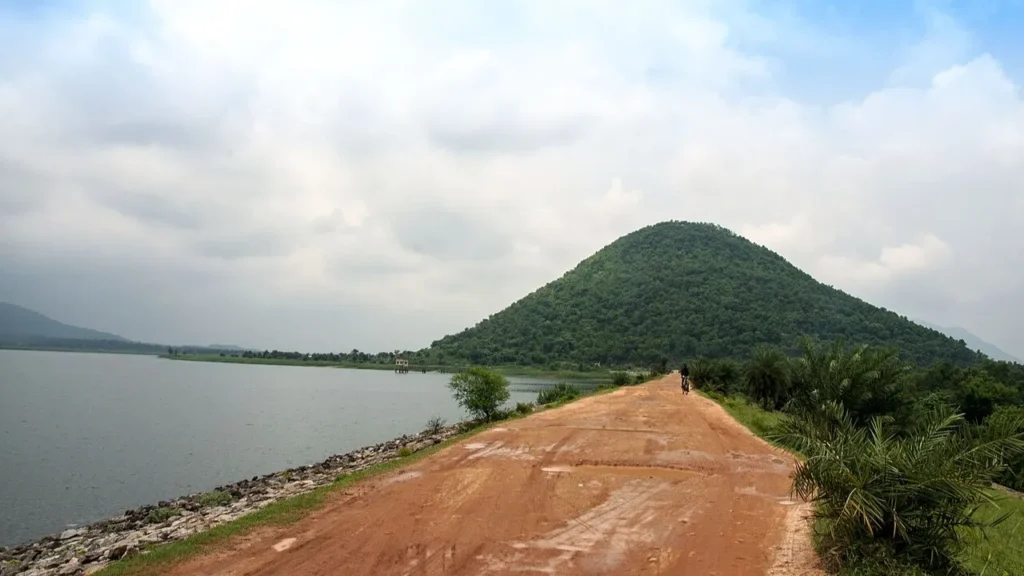 A scenic view of Baranti Lake and Baranti Hill in Purulia, West Bengal, featuring a red dirt road alongside a calm reservoir under a cloudy sky, surrounded by lush green forests.