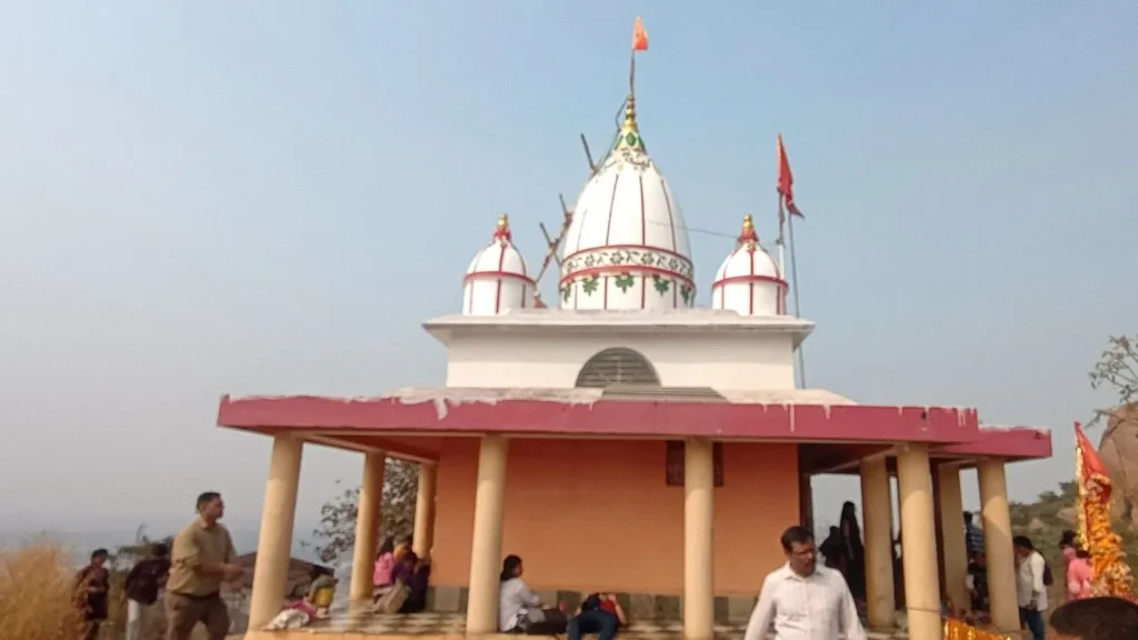The Chandi Mata Temple at the summit of Joychandi Pahar in Purulia, West Bengal, featuring white domes with red accents, orange flags, and devotees resting in the open-air pavilion against a clear sky.