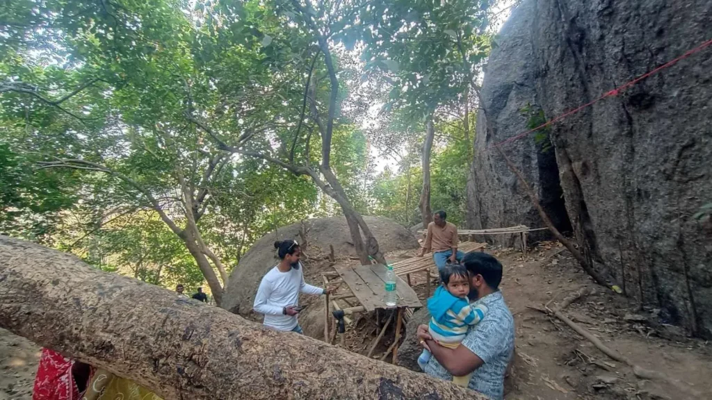 A rustic outdoor rest area at Joychandi Pahar in Purulia, featuring people sitting at wooden tables under a canopy of trees next to a large granite rock formation.