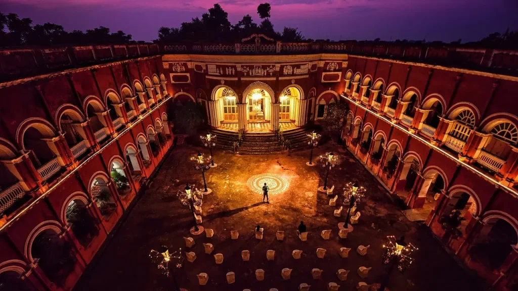 An aerial nighttime view of the illuminated inner courtyard of Itachuna Rajbari heritage home in West Bengal, featuring red-ochre walls and classical arches under a twilight sky.