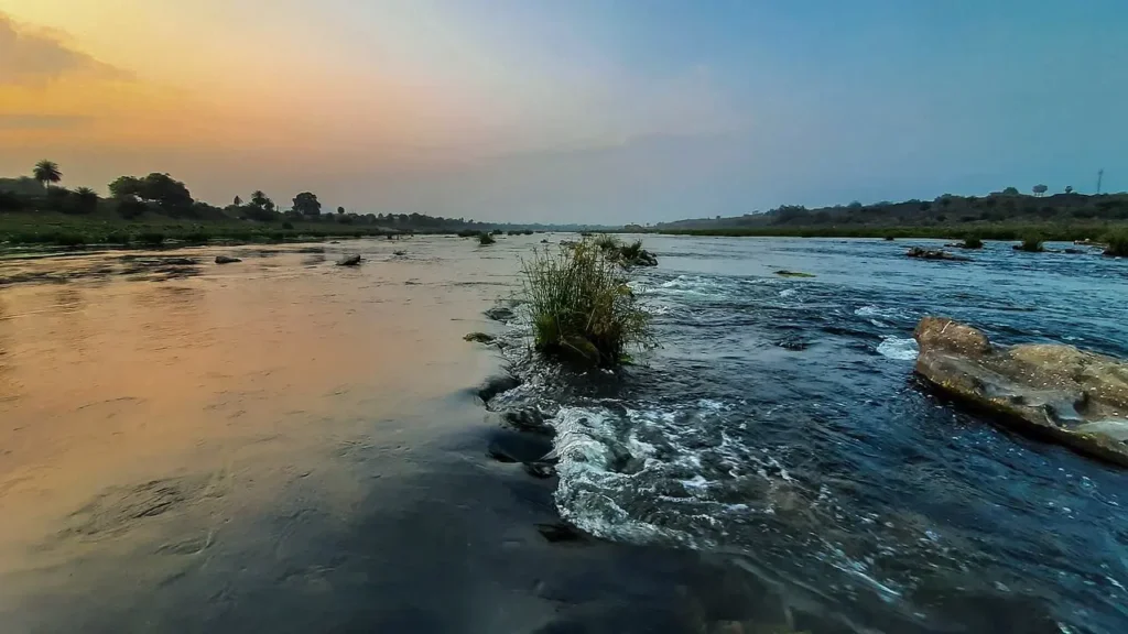 A scenic wide-angle view of the Subarnarekha River in Ghatshila at sunset, featuring flowing water over rocks and a vibrant golden and blue sky.