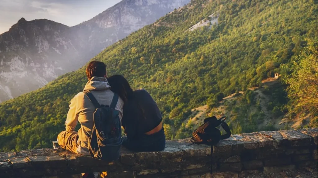 A romantic couple sitting on a rustic stone wall, overlooking a vast, lush mountain valley during a golden sunset.