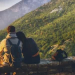 A romantic couple sitting on a rustic stone wall, overlooking a vast, lush mountain valley during a golden sunset.