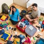 A stressed Bengali couple sitting on a wooden floor surrounded by scattered colorful clothes, trying to pack a red suitcase using the 5-4-3-2-1 travel packing hack with a Travel Packing Checklist.