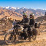 A man and a woman in full riding gear (helmets, jackets, pants) are on a Royal Enfield Himalayan adventure motorcycle heavily loaded with hard panniers, a top box, and a tall rear camera pole with a 360-degree camera. They are on a dusty mountain track in an arid Himalayan landscape with traditional village buildings in the background and snow-capped peaks in the distance. The man is riding, and the woman is the pillion passenger.
