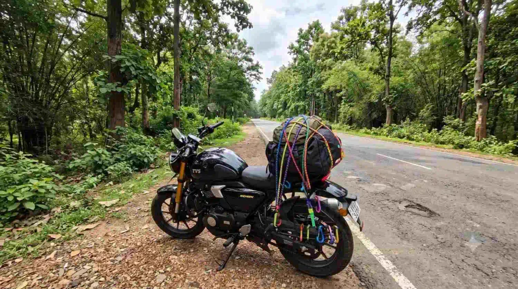 A Lightning Black TVS Ronin parked on a rural asphalt road shoulder, rear seat loaded with touring luggage secured by colorful bungee cords against a dense green forest backdrop.