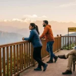 A woman and a man stand at a wooden railing, looking out at a vast, snow-capped Himalayan mountain range during golden hour. A sign nearby reads "GHOOM STATION". A third man sits on a bench to the right, reading a book. The sky is a warm gradient of orange and pink