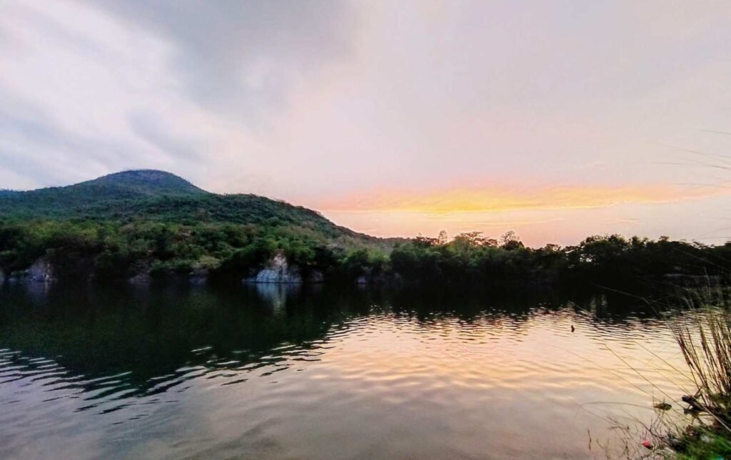 Calm waters of Blue Lake in Balasore at dusk, reflecting a warm gradient of golden-yellow and soft pink from the sunset sky, framed by rolling green hills and rocky shoreline greenery.